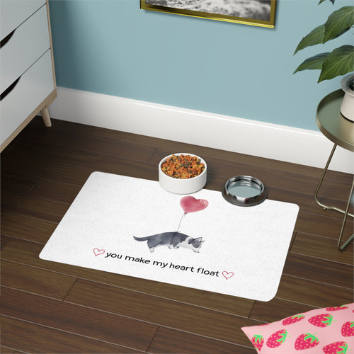 White mat that says "you make my heart float" with a floating dog on a wooden floor, accompanied by a bowl of food and a water dish.