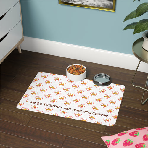 Pet mat with paw, hearts, and mac and cheese pattern on a wooden floor, accompanied by a bowl of food and a water dish.