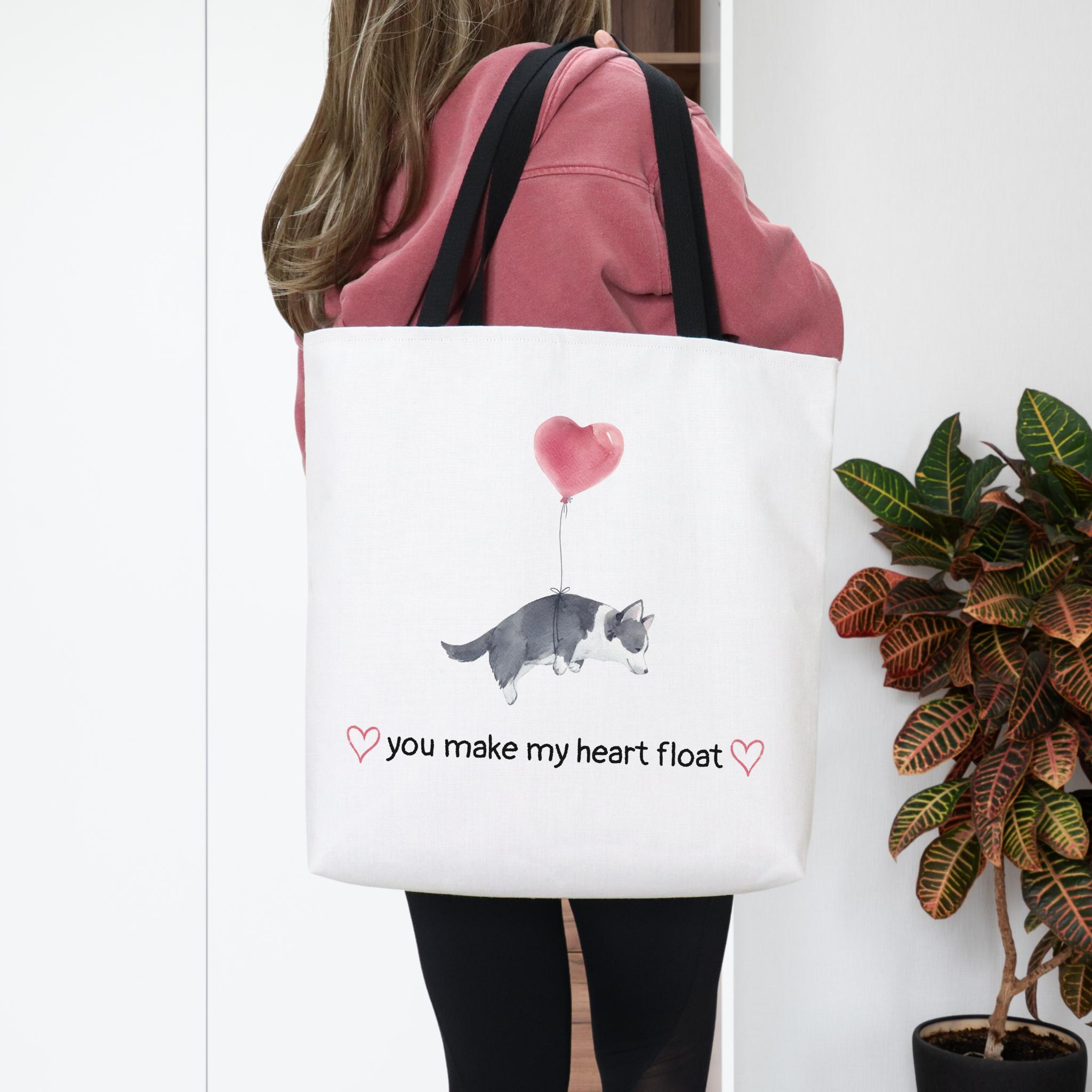 Person holding a tote bag with a design of a dog with a heart balloon and text 'you make my heart float'.
