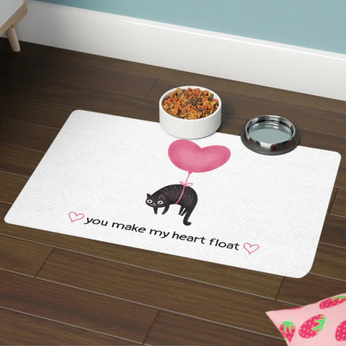 Pet mat with a floating black cat and heart design and text on a wooden floor.